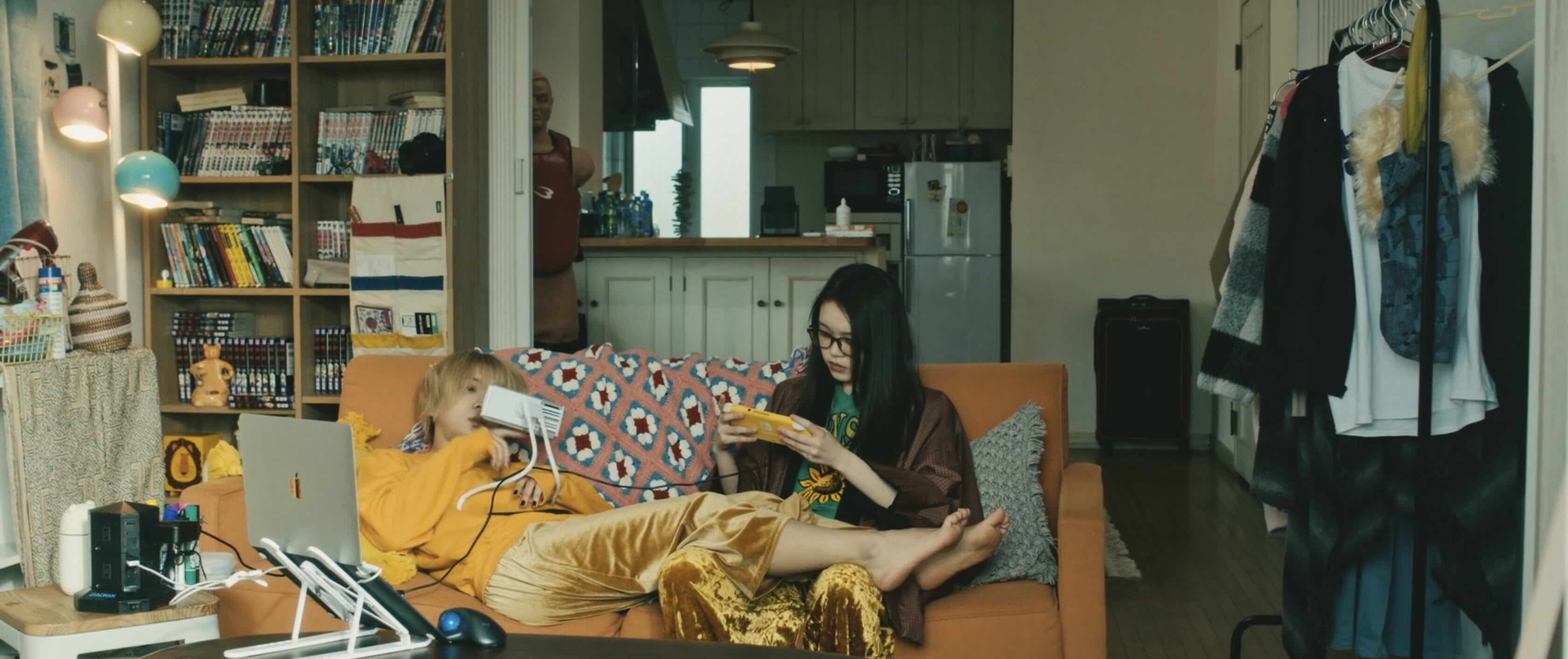 Two bored looking Japanese teenage girls slumped on a sofa, one playing a Nintendo Switch, the other watching her mobile phone.