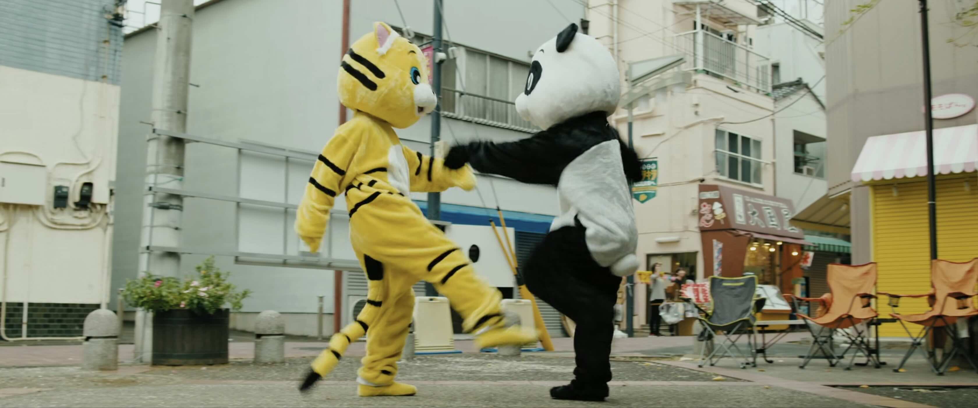 Two people in furry mascot suits, one as a tiger, the other as a Panda. The tiger is kicking the panda.
