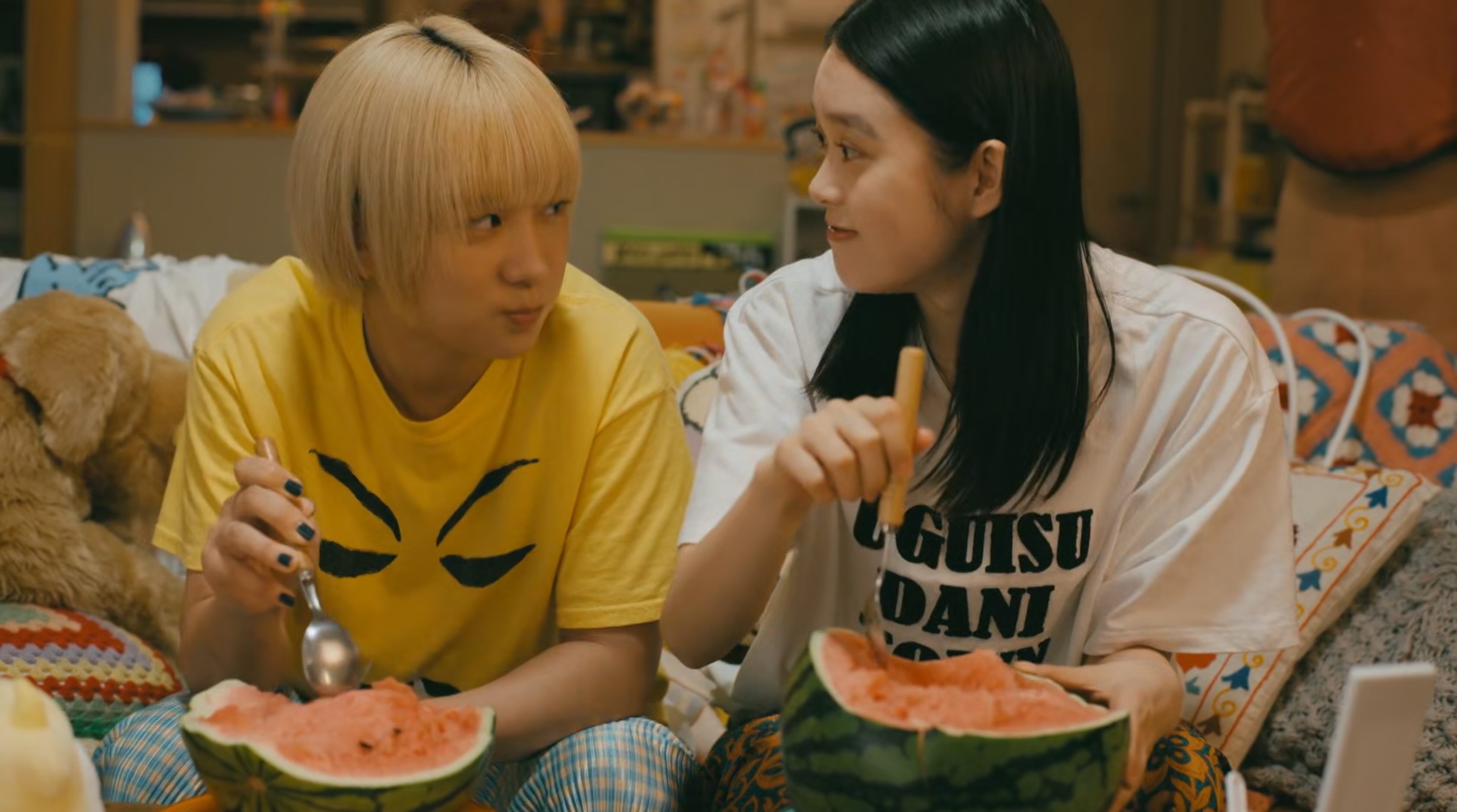 Two Japanese teenagers sat on a sofa eating a large melon with spoons. The one on the left is blonde, the one on the right is brunette and has a goofy expression. They're both looking into each other's eyes.