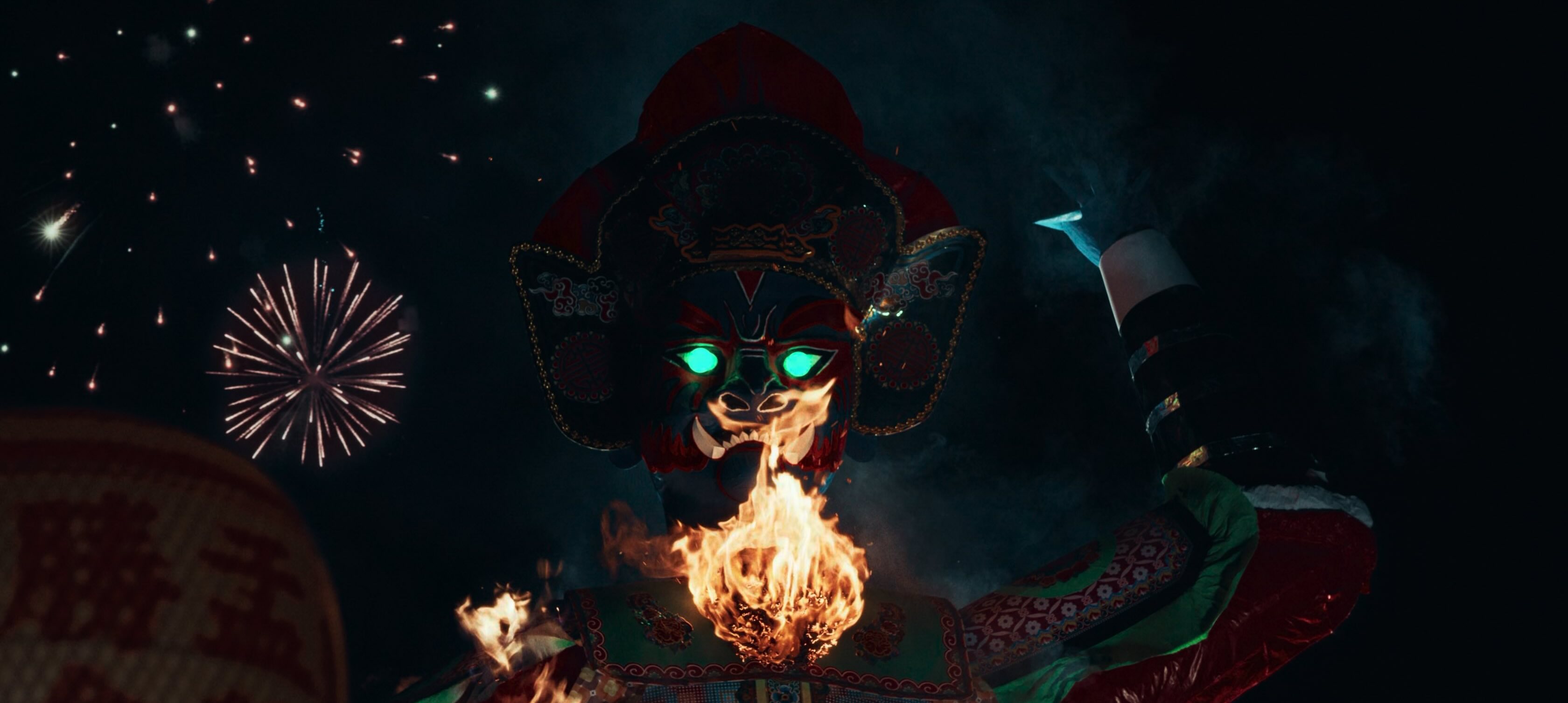 A nighttime scene of a huge chinese effigy. It's chest is on fire, fireworks explode in the background