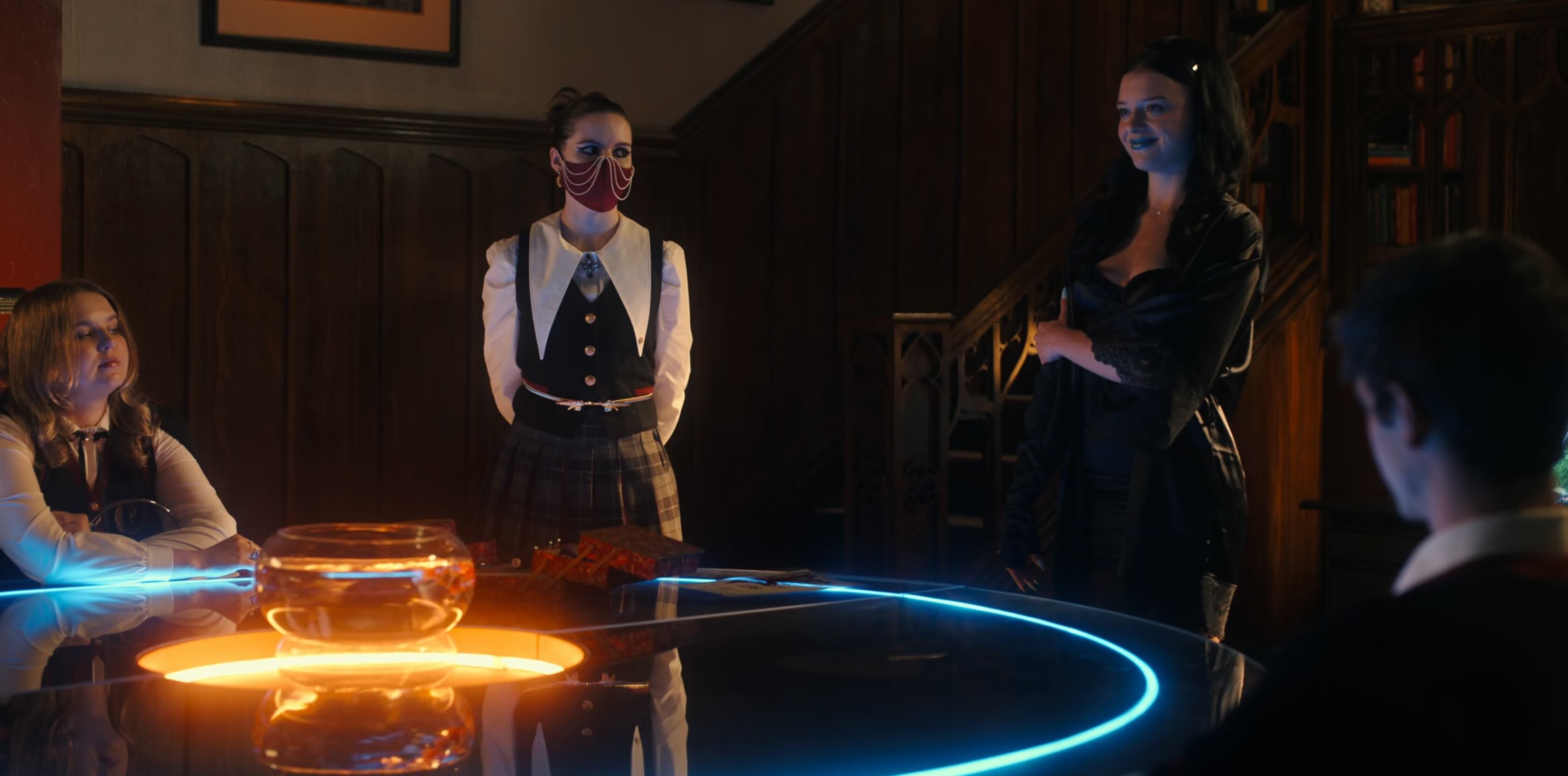A glass round table, highlighted in blue and orange LED lights. Around which stand several school students. The most prominent is wearing a red covid mask with gold chains draped on it, a tartan skirt and blouse/waistcoat combination with absolutely huge draped collars.