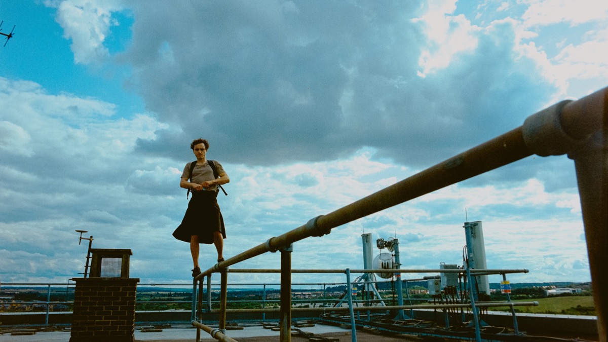A man in a long kilt stands on scaffolding on top of a building. An overcast sky takes up most of the picture, stretching to the far away, flat horizon