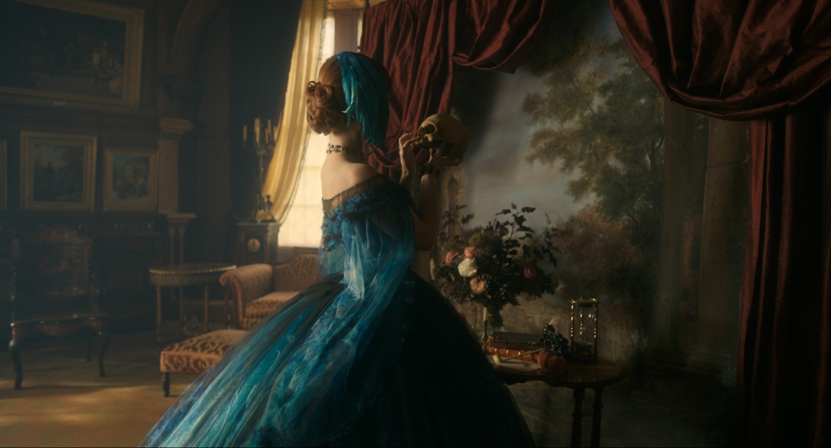 An absolutely sumptuous still shot of a woman in a nineteenth century, azure blue dress, layered with chiffon. She's holding a human skull and looking at it intensely. The room is filled with baroque artwork, furniture and curtains.