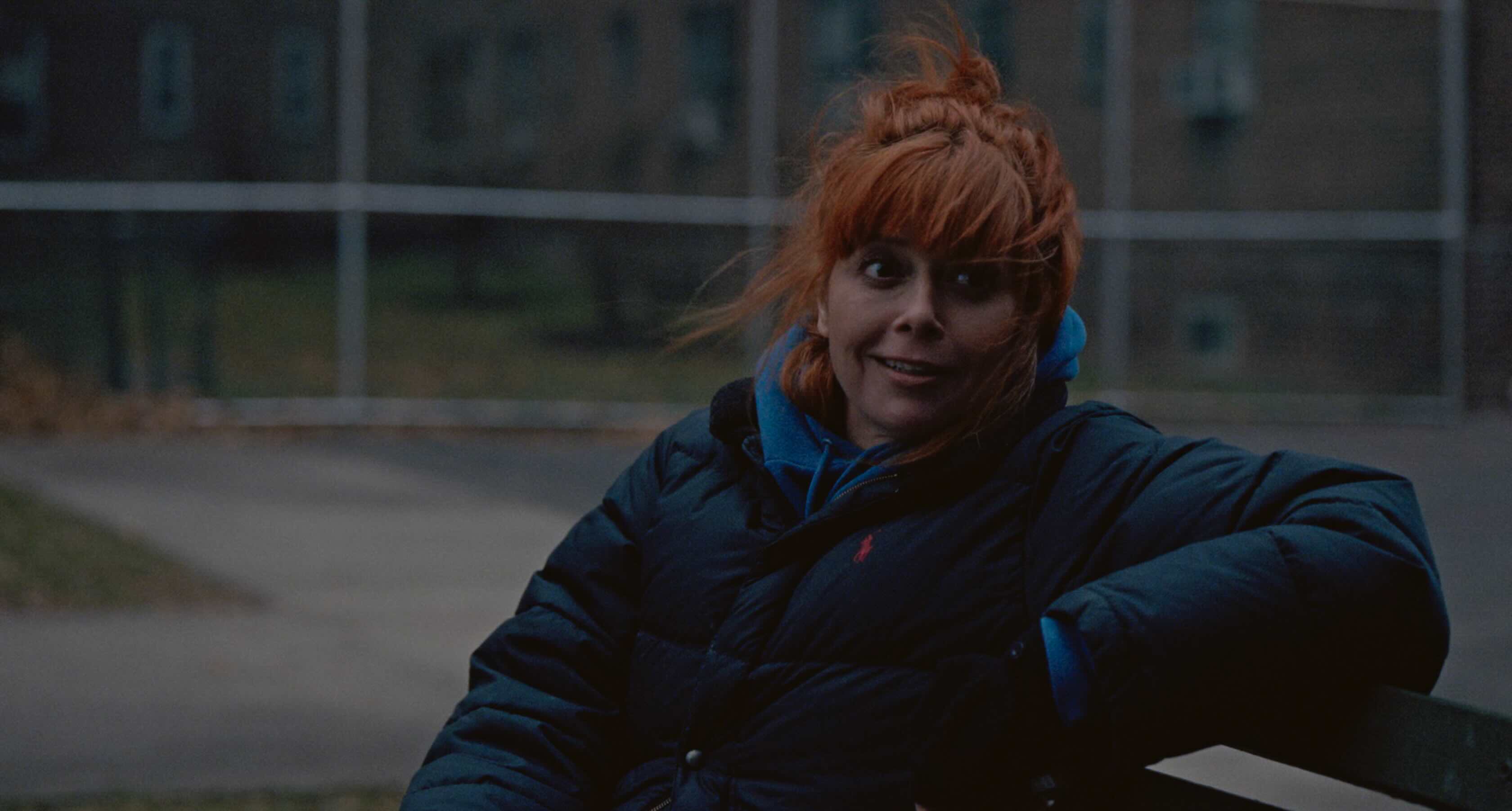 Natasha Lyon, with flame red hair and a navy puffa jacket, sitting on a park bench
