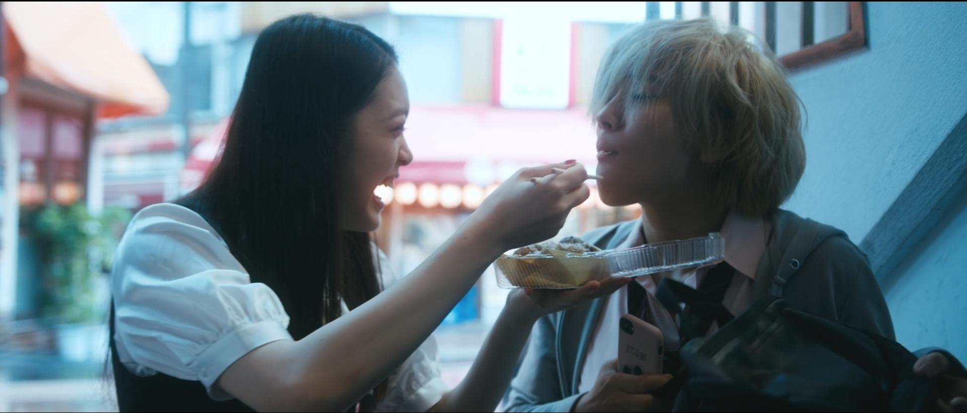 Two Japanese teenage girls. The long haired brunette on the left is using chopsticks to feed sushi to the short haired blonde on the right.