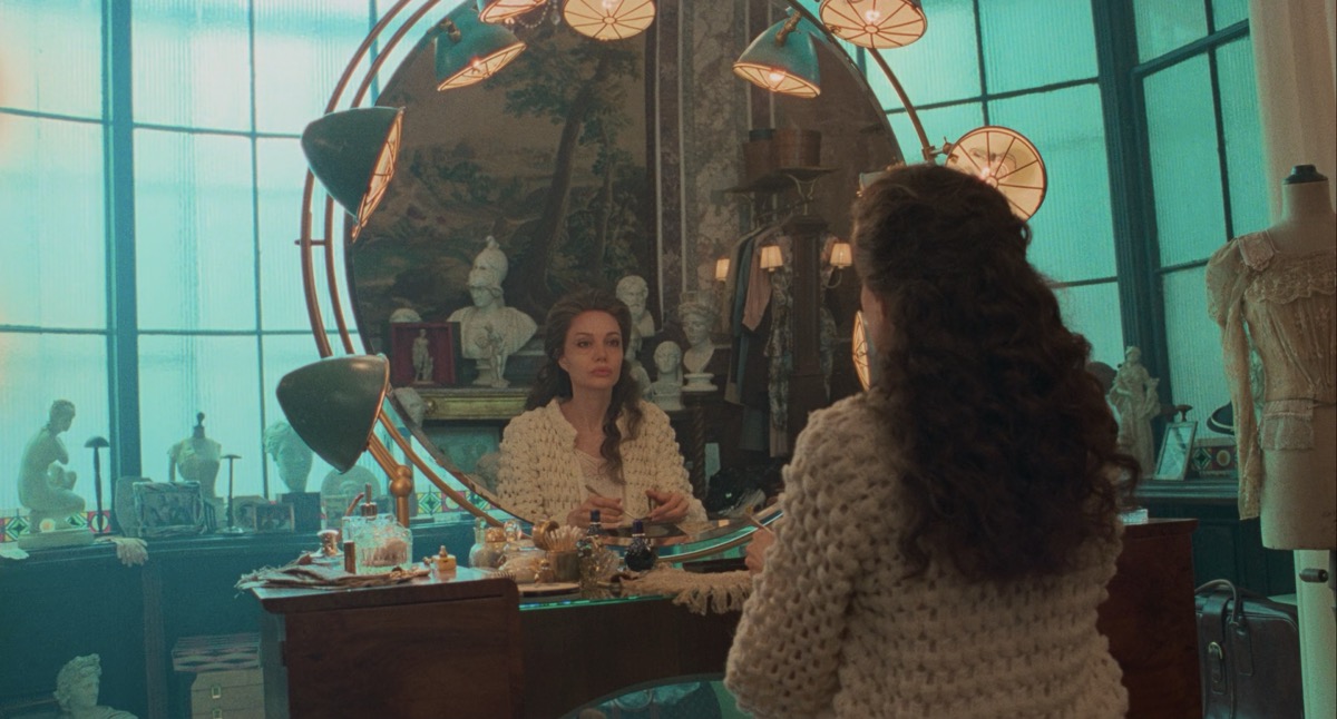 Angelina Jolie looking into a large round mirror, lit with large round lamps. In the mirror we can see a series of classical marble busts behind her