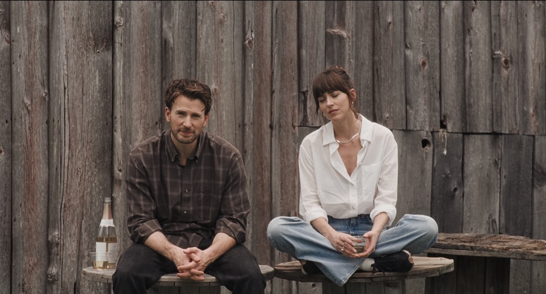 Chris Evans and Dakota Johnson looking out at the camera, sat in front of a wooden fence