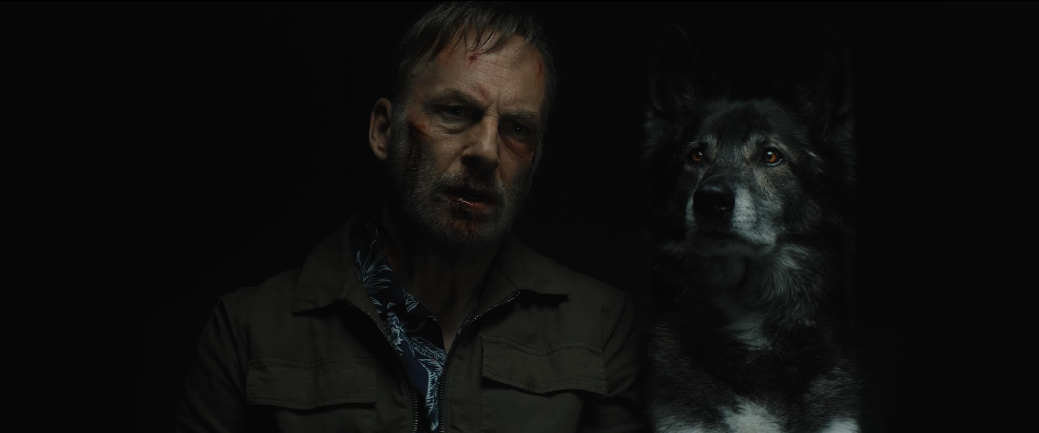 A middle aged man, badly beaten up, looking very morose sat next to a black and grey dog looking very alert and attentive