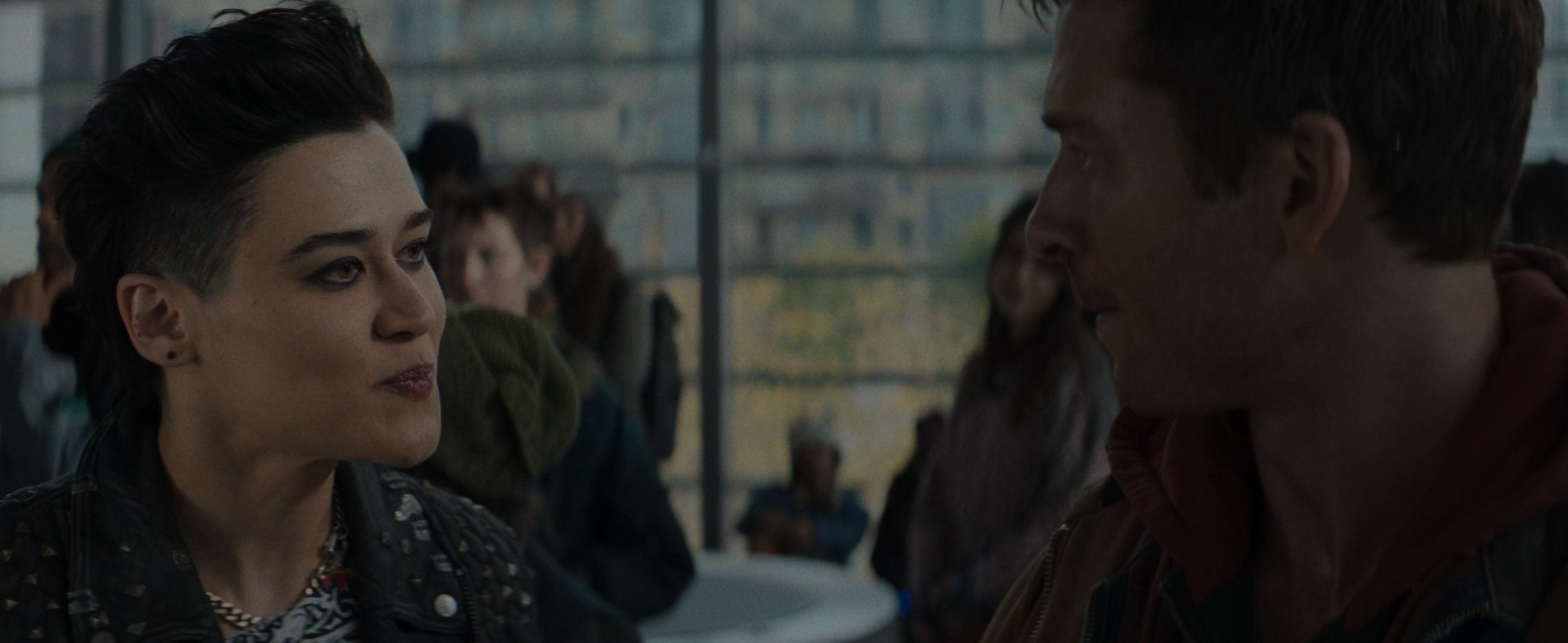 A punky looking white woman with short black hair, sharp black eyeliner and a studded jacket talking to a square jawed white guy.