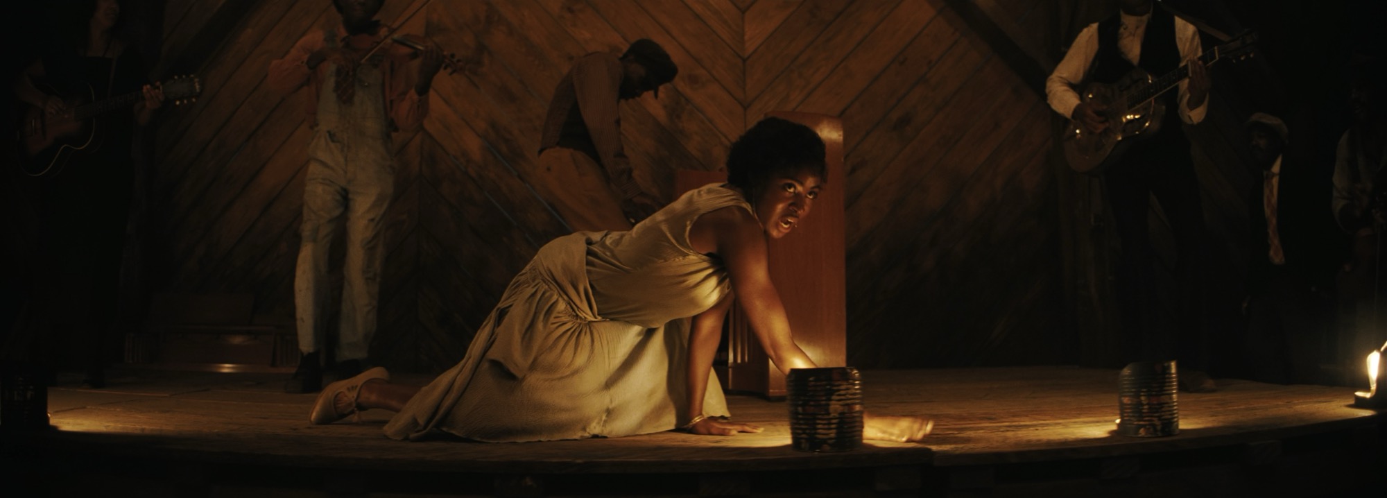 a woman in a dress crawling on a stage, singing. Behind her a band plays a fiddle, piano and guitar