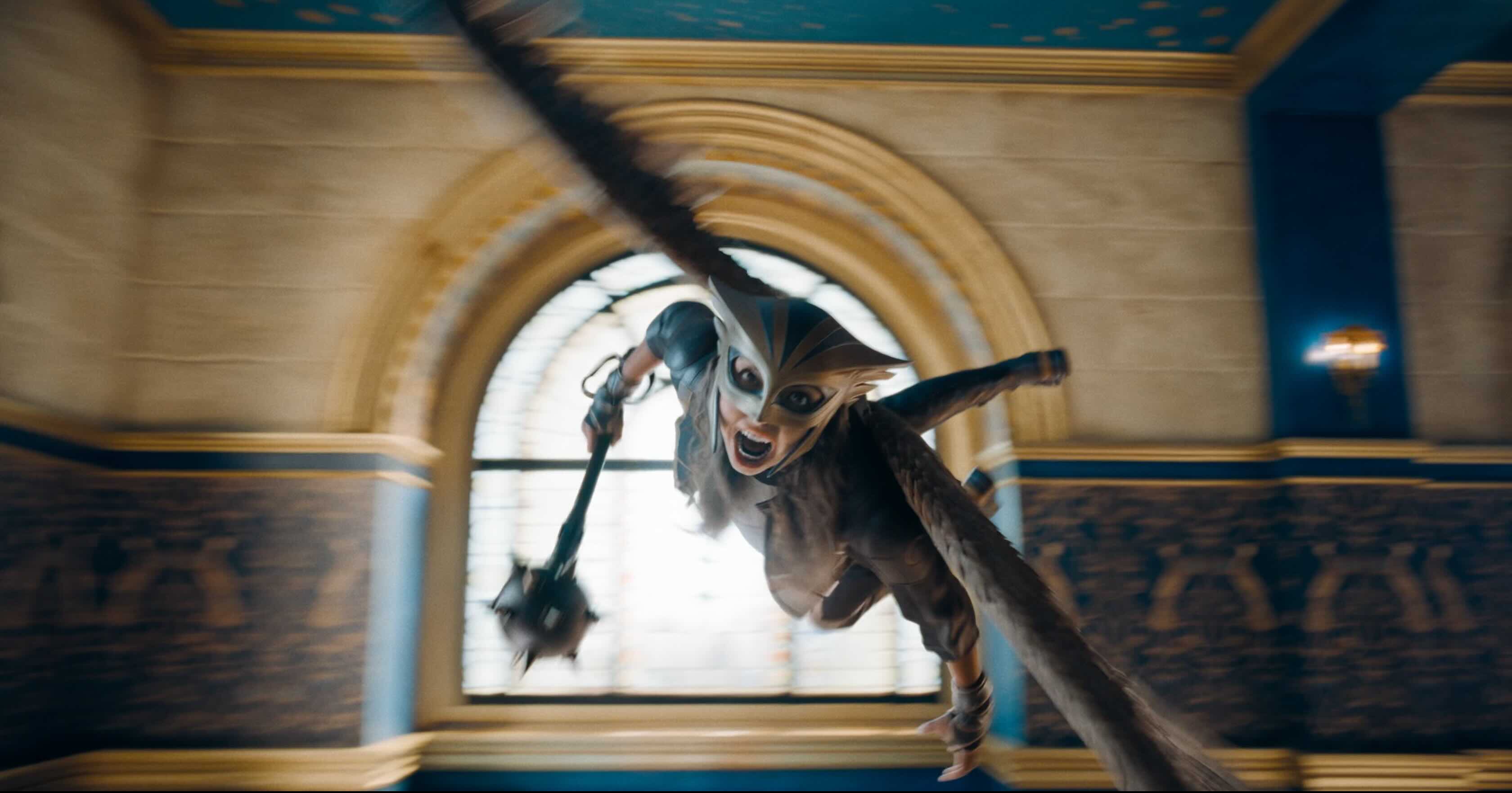 A young, female superhero with wings and a mace flies at the camera screaming