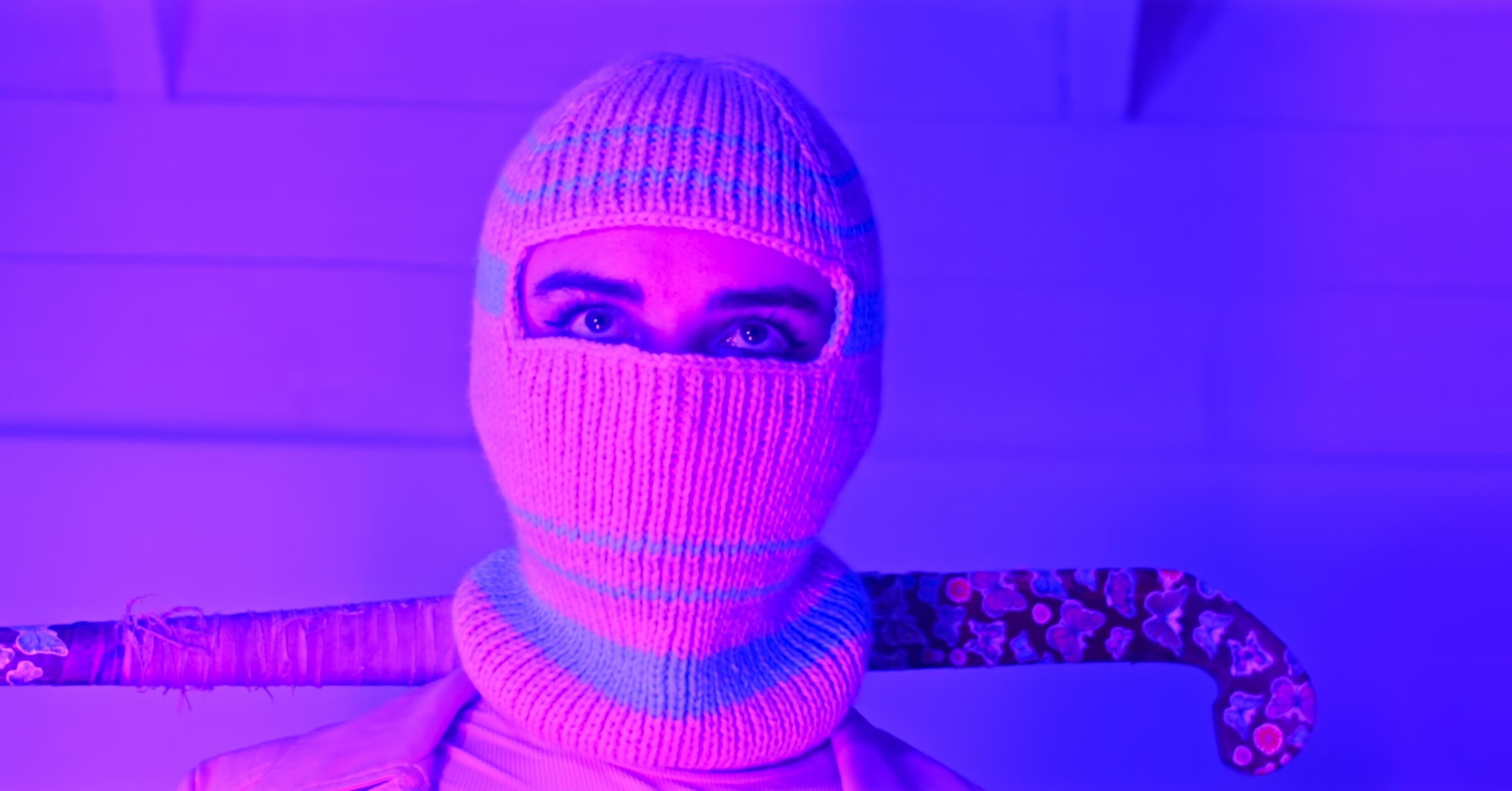 A woman wearing a white balaclava, holding a hockey stick. The lighting douses everything in a pink glow.