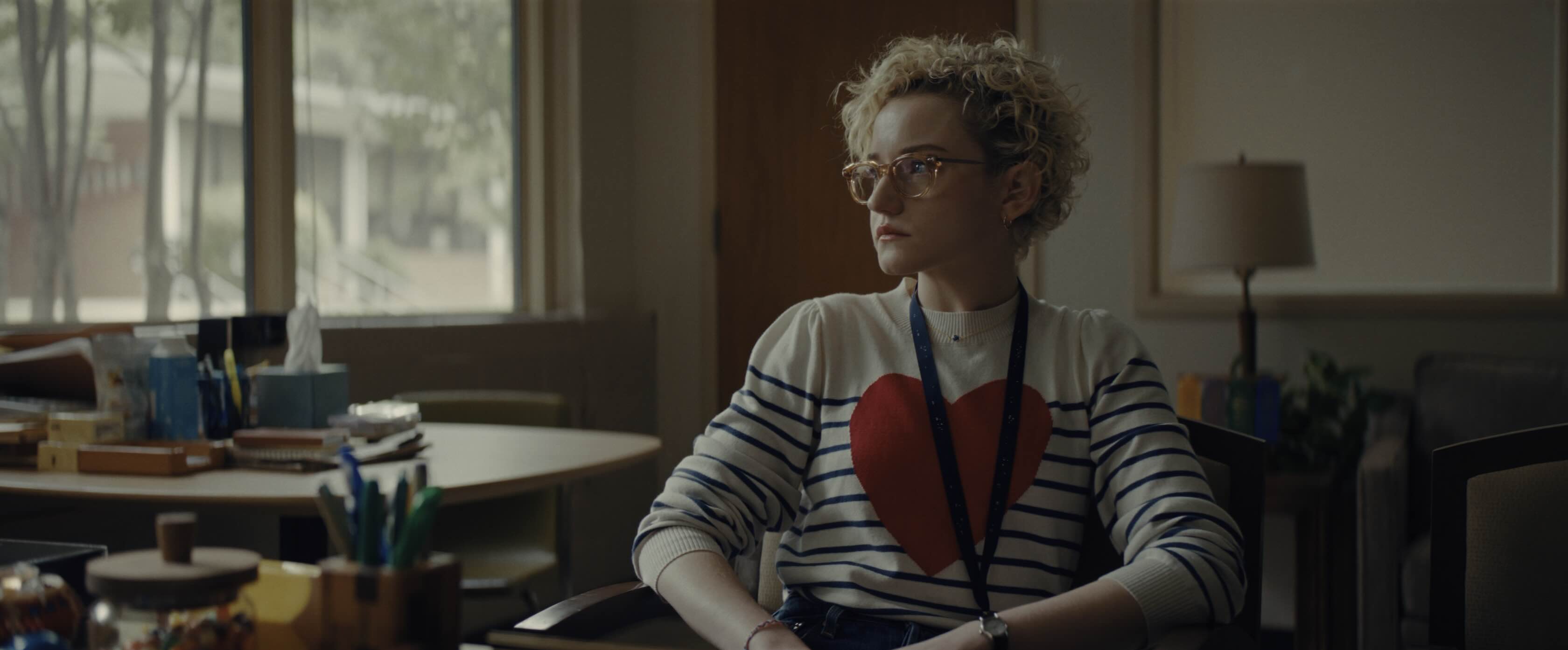 A woman with short, curly blonde hair, wearing a jumper with a heart on it, sitting in a primary school class room.