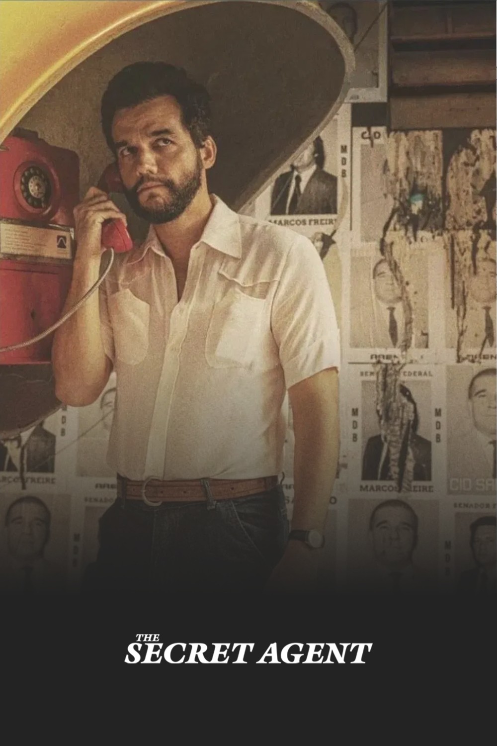 Poster for the film The Secret Agent. A Handsome Brazilian man stands in a yellow telephone kiosk. He has a well trimmed beard, a crisp white, short sleeved shirt and is looking cautiously to the side. The background is wall plastered with faded ripped poster of mugshots.