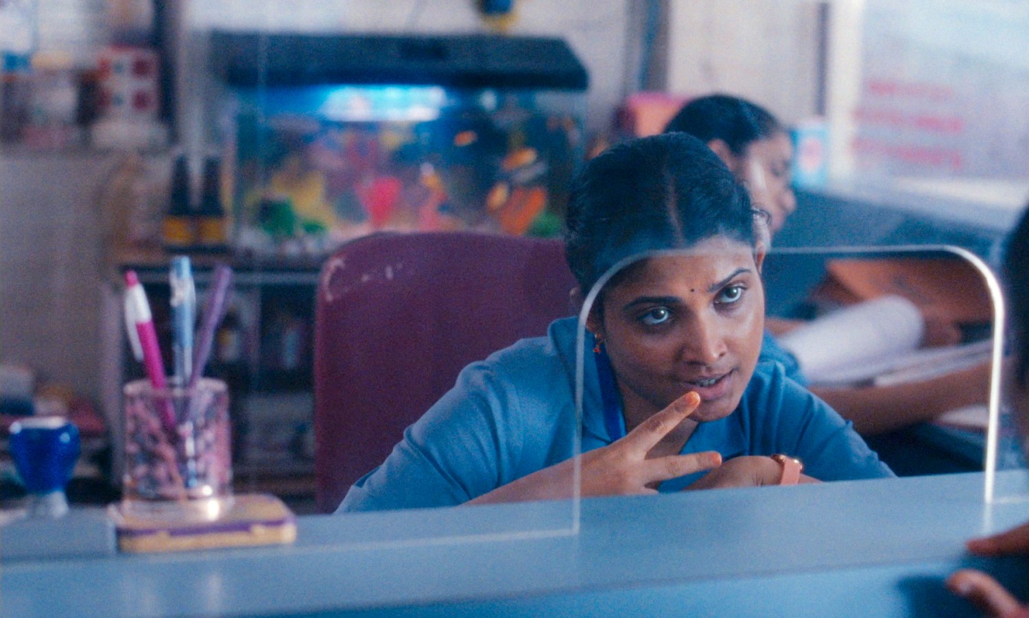 A young Indian Nurse sits behind a glass partition in a busy doctor's surgery. Behind her are cluttered desks and a colourful fish tank. She's talking to a young mother, just off camera, and making the 'snipping scissors' action with her fingers.
