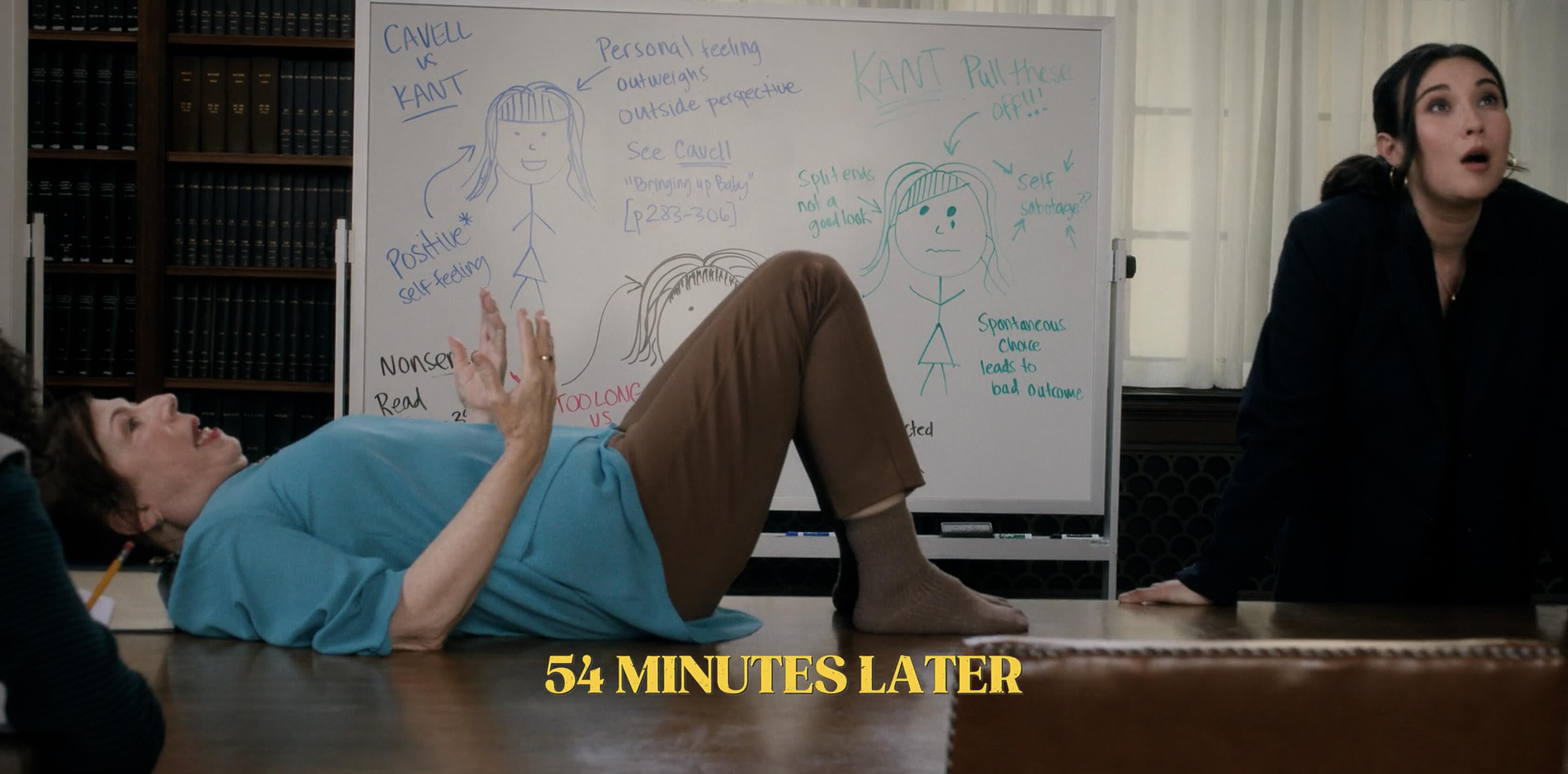 An older woman with no shoes lays on a big wooden desk, gesturing with her arms. Behind her is a whiteboard with a mixture of philosophical points written on it and cartoon stick figures. A younger woman in a business suit has her hands clasping the desk and a look of despair.