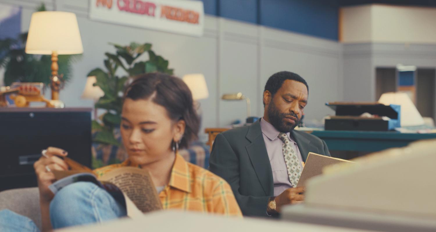 A young white woman, dresses in a casual yellow check shirt and jeans, and a middle aged black man, dressed in a smart suit, both sit at a desk in a bland furniture store. Both are intently reading, not paying attention to each other.