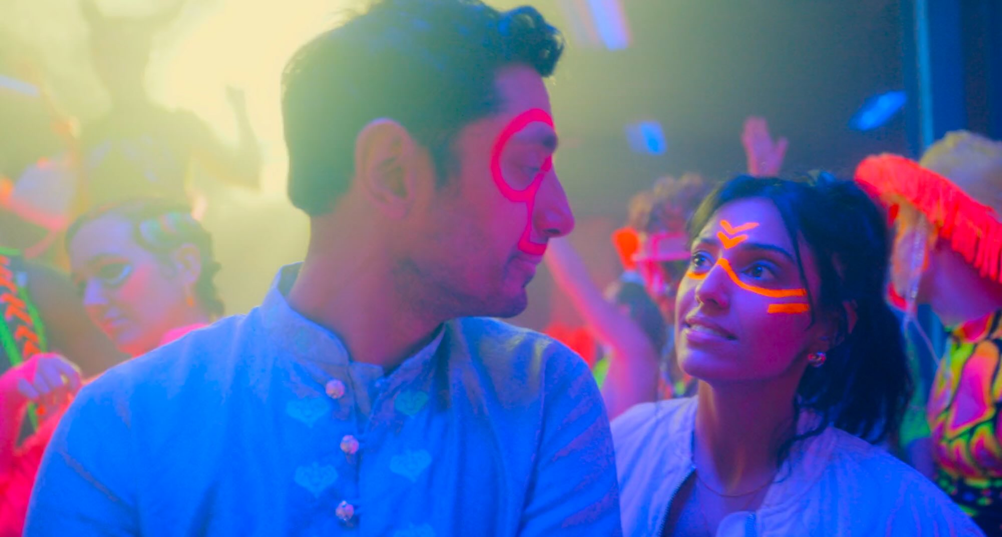 A club scene. It's darkly lit but everyone is wearing neon, glow in the dark clothes and jewellery. The two characters in the foreground are a young Asian man and woman, faces covered in lines of brightly glowing paint, having a conversation.
