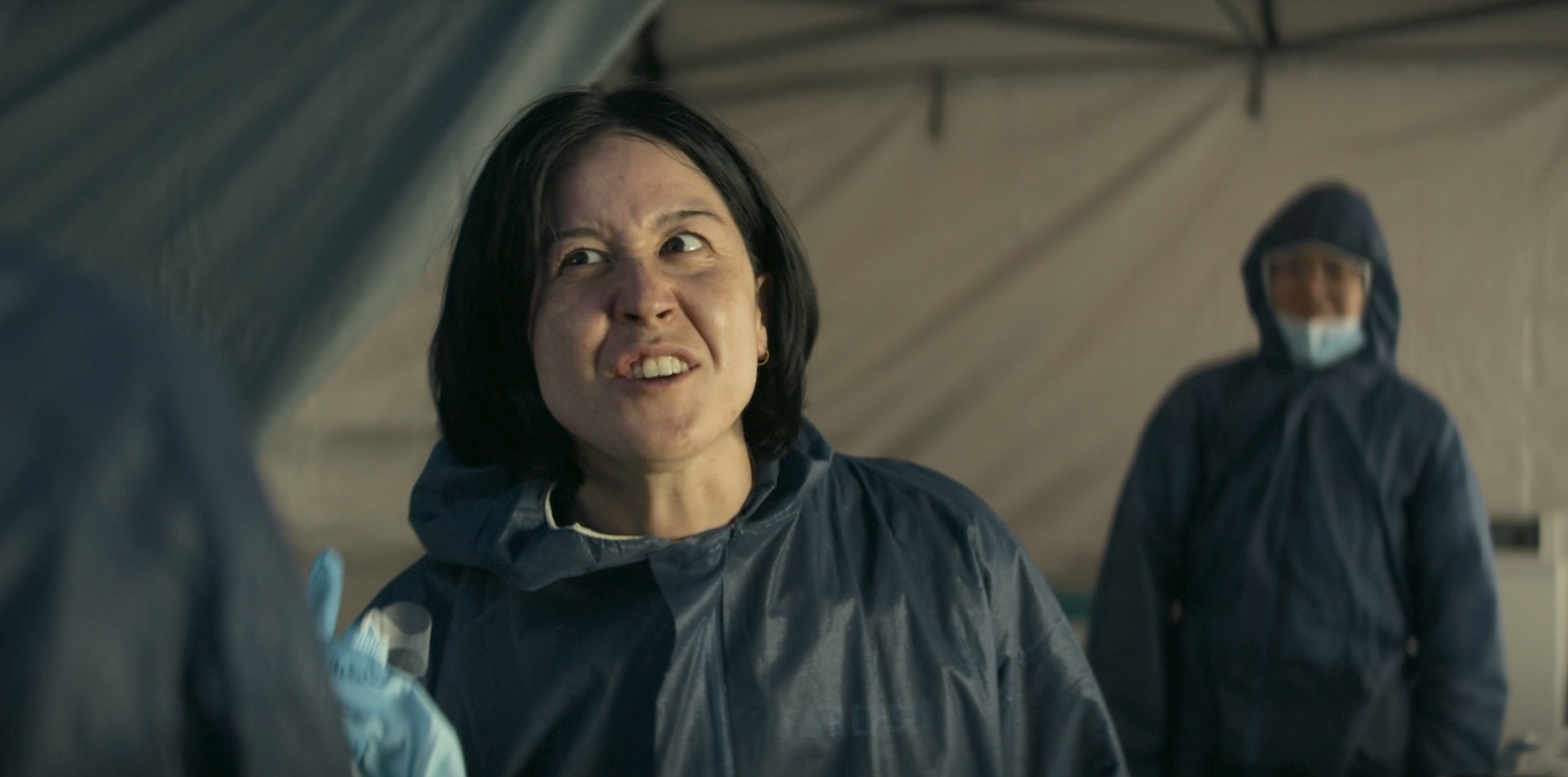 A small, timid, mousey young woman wearing a blue, forensic investigation suit. She's yelling foul obscenities and insults at someone just off camera. Behind her is a small child, also in full forensic gear.