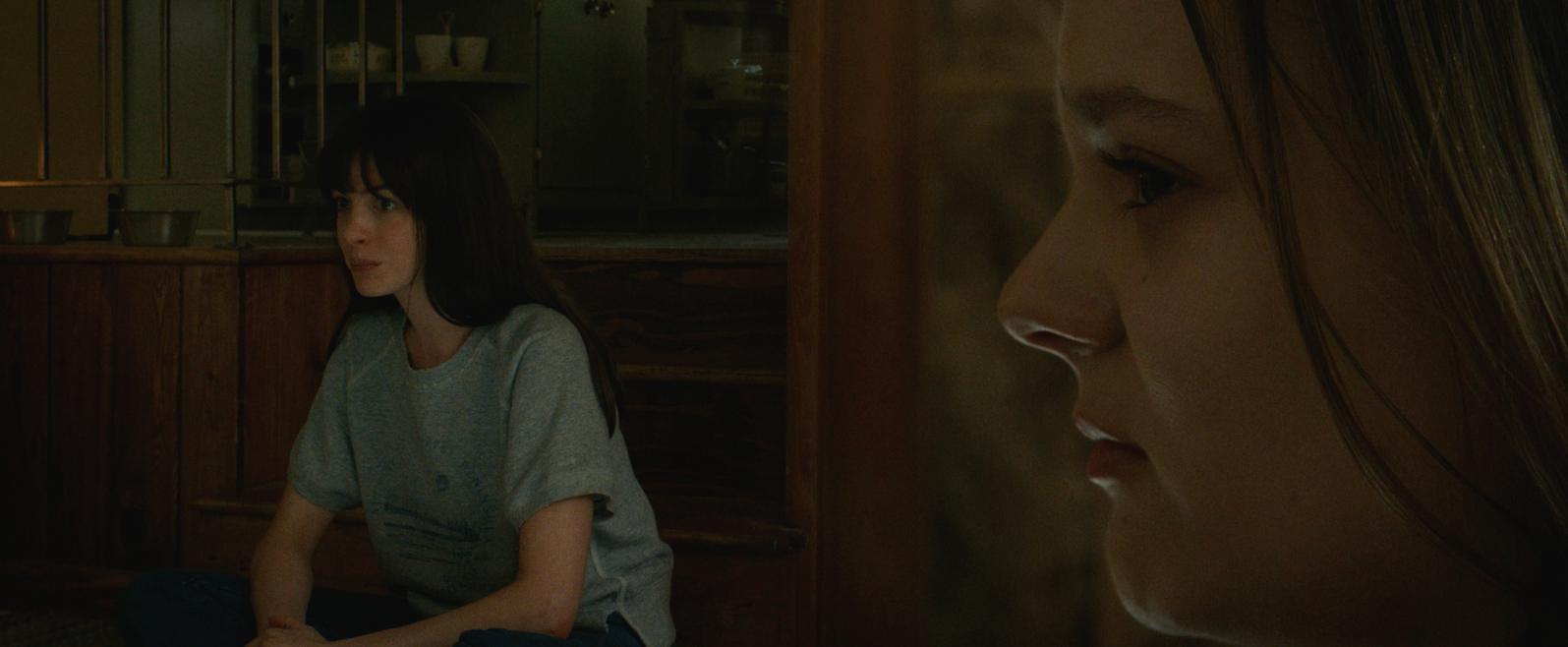 A dark interior of a home with late seventies decor. On the left half of the picture a dark haired, middle aged woman is sitting cross legged. The right half of the picture is taken up by a very close head shot of a blond teenage girl looking at the other woman.