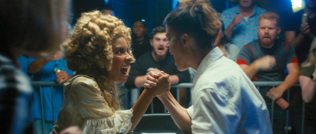 Two women engaged in an arm wrestling match, screaming at each other in effort. One is dressed like Marie Antoinette with a gold filagree dress with cuffs and wearing a big golden curled wig, the other is dressed like a baker in a crisp white shirt. In the background is a baying and cheering crowd.