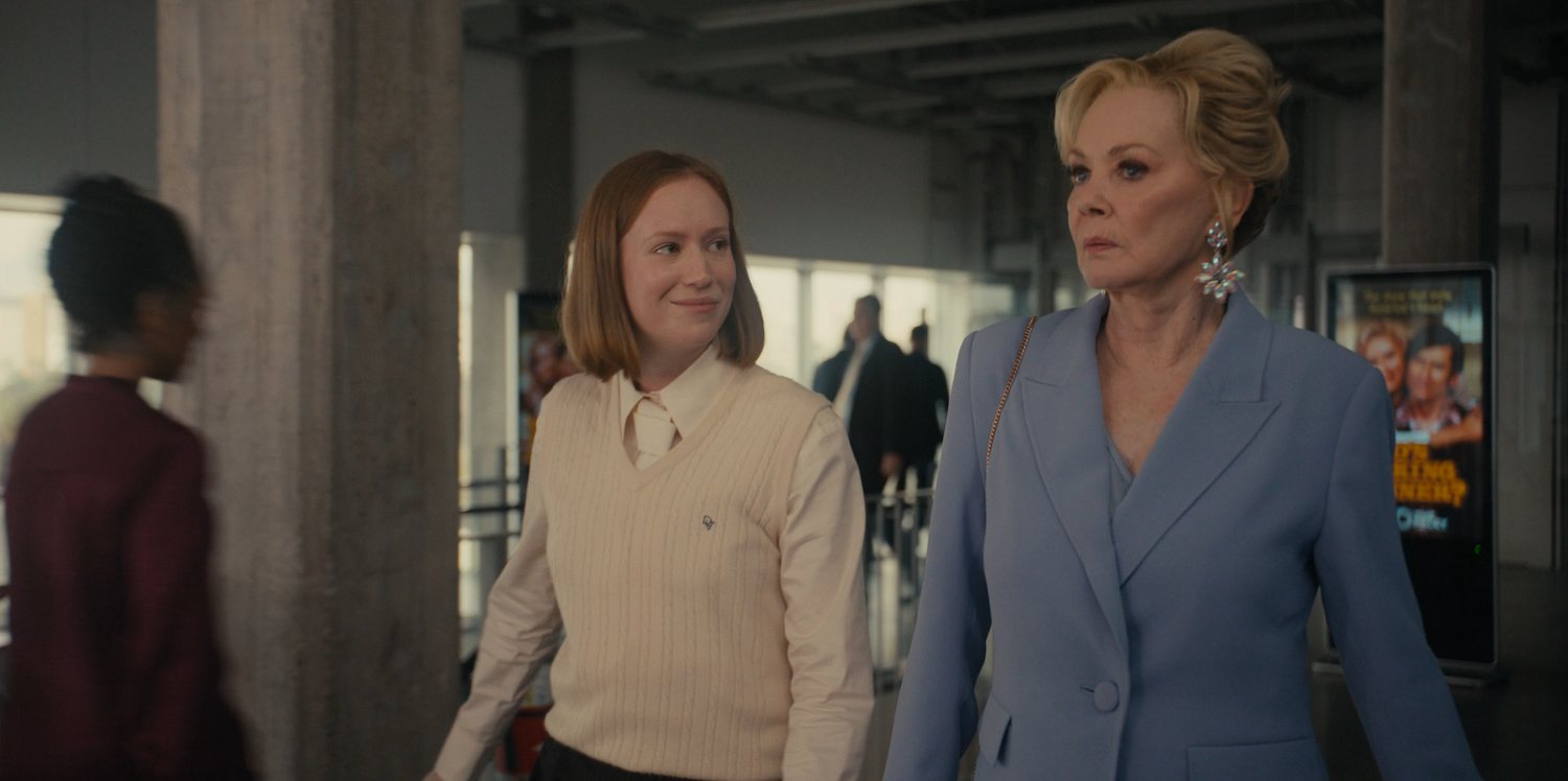 The same two women from the poster walking side be side in a conference space. The younger woman is wearing a cream, sleeveless sweater over a cream blouse with a cream necktie. She looks a little like a very formal cricketer. She's looking at the older woman, amused. The older woman is wearing a dusty blue suit and big silver flower earrings. She does not look amused.
