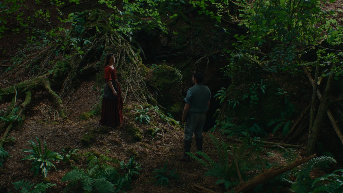 A high camera shot looking down at an overgrown, lush green forest. A woman in a red dress and a man in a light blue outfit are standing some distance apart, stanced slightly awkwardly, looking at each other.