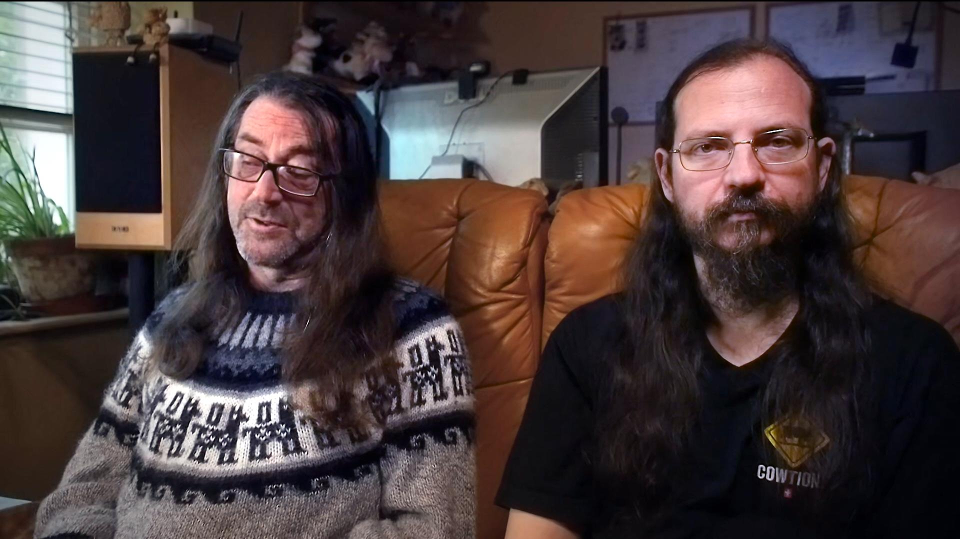 Two men, both with beards, glasses and long brown hair, sit side by side on a brown leather sofa.