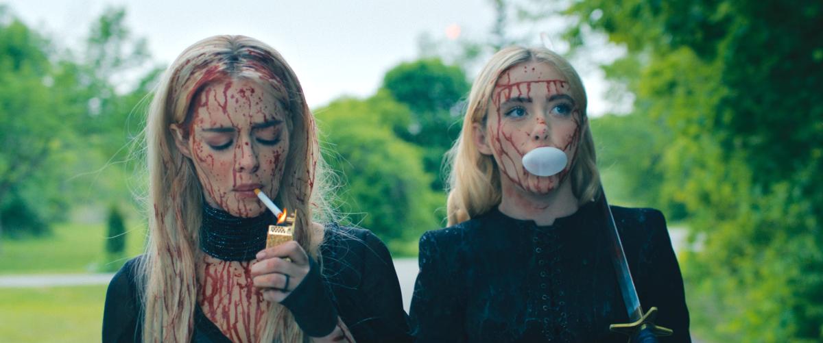 Two young blonde women wearing gothic black dresses, faces covered in splatters of blood. One is lighting a cigarette, the other is blowing a bubblegum bubble and has a sword slung over her shoulder.