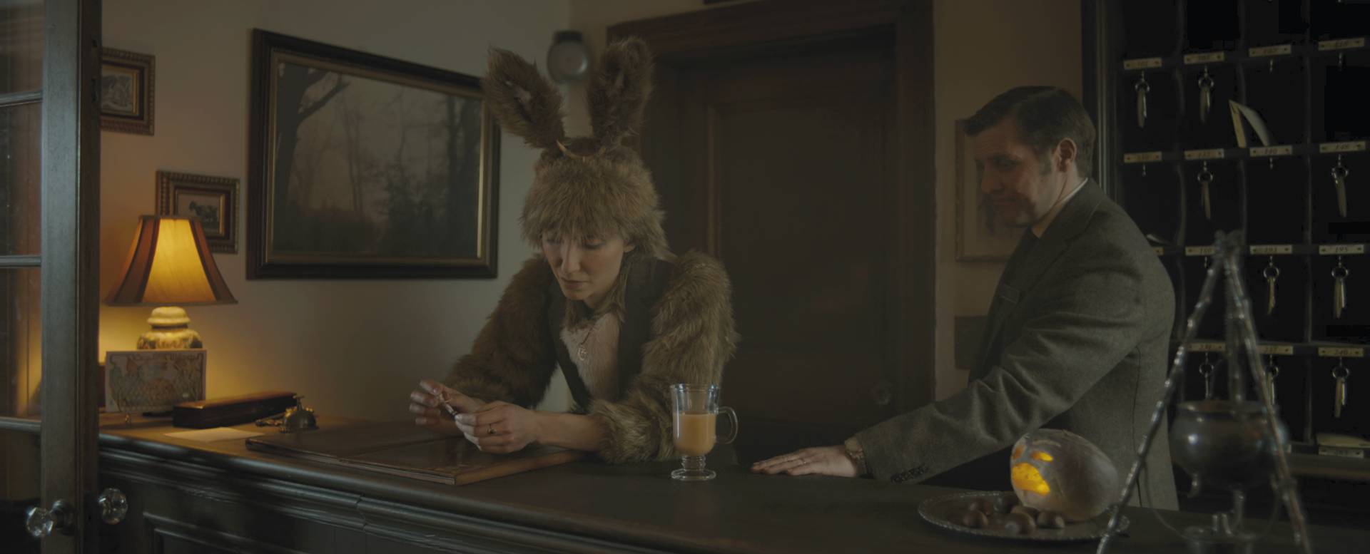 A man and a woman stood behind the reception desk at an old fashioned, dingy, Irish hotel. The scene is dimly lit by a dull table lamp of to the side. She is wearing a shaggy, brown rabbit costume. He is wearing a dull, grey suit.