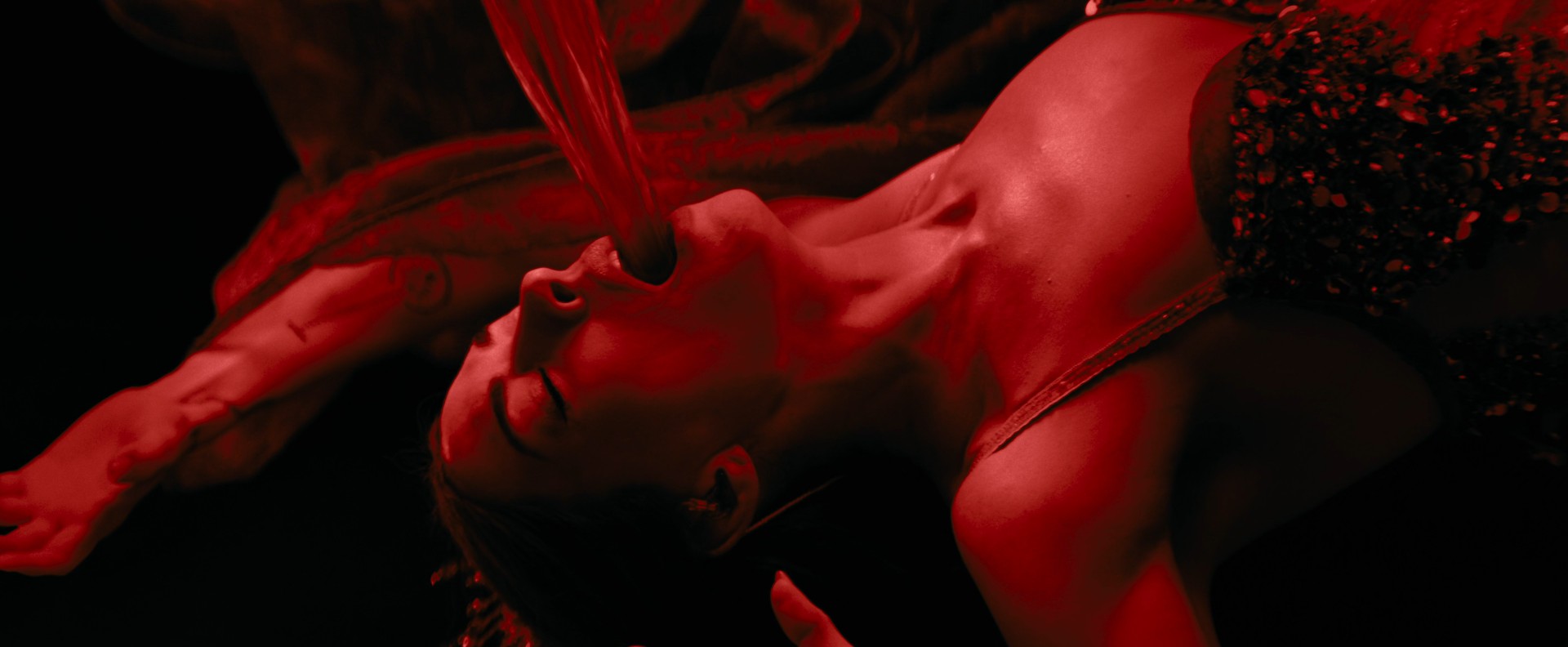 A woman is lying on her back, hands stretched above her head, back arched. A stream of thin fabric is pouring either out of or into her mouth. The whole image is shot in a black and red colour scheme.