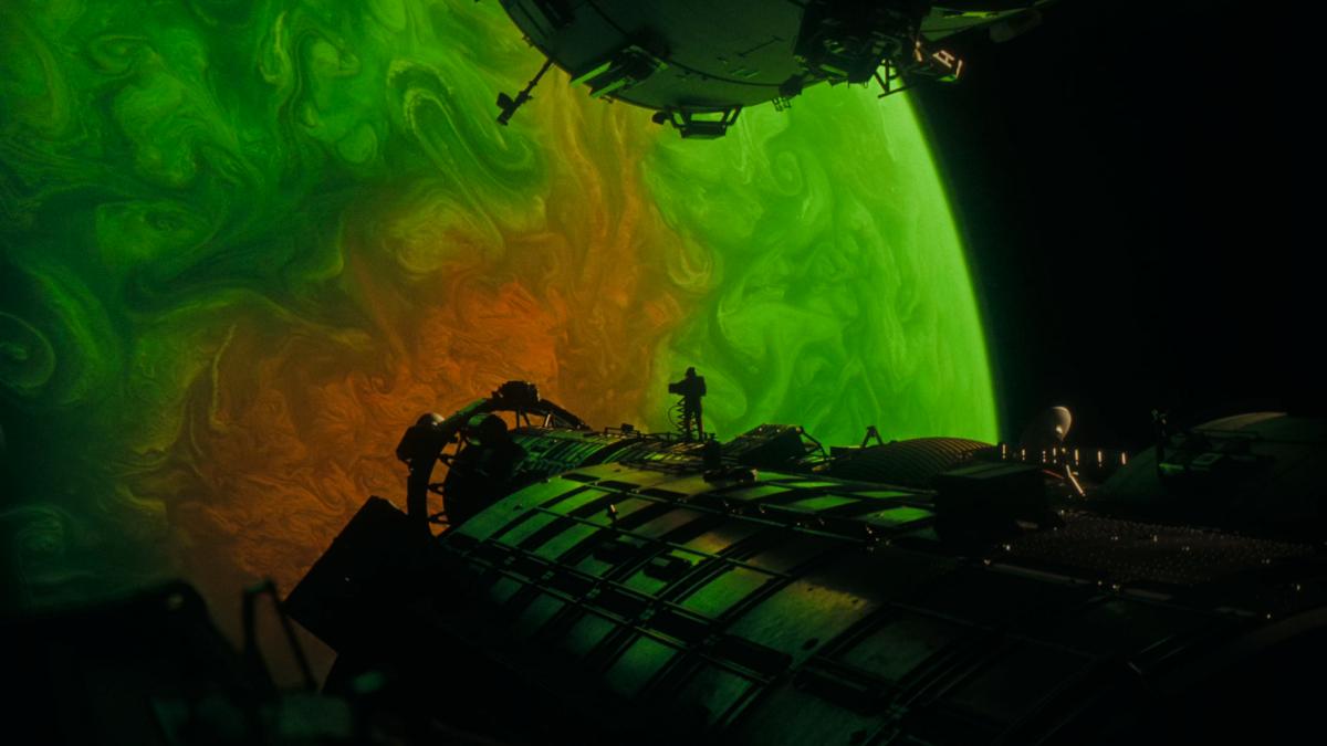 A photo, taken from the point of view of someone standing on a space ship. Part of the ship loom at the sides of the shot and we can see an astronaut standing on the ship in the distance. Everything is coloured in a greenish hue that is being given off by the giant, green and orange, gas planet which completely dominates the view.