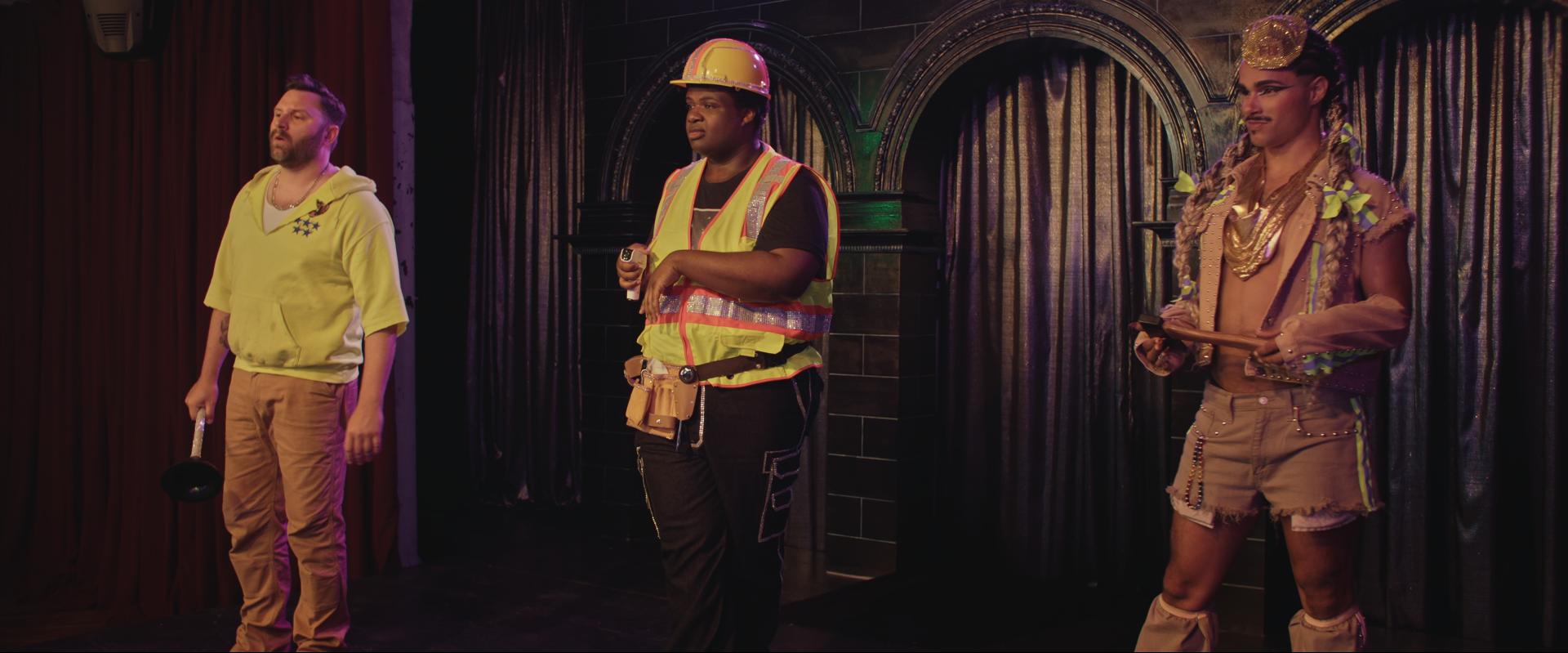3 male or non-binary people standing on a stage. Dressed as very, very camp emergency workers: a plumber, a construction worker and a fire fighter.