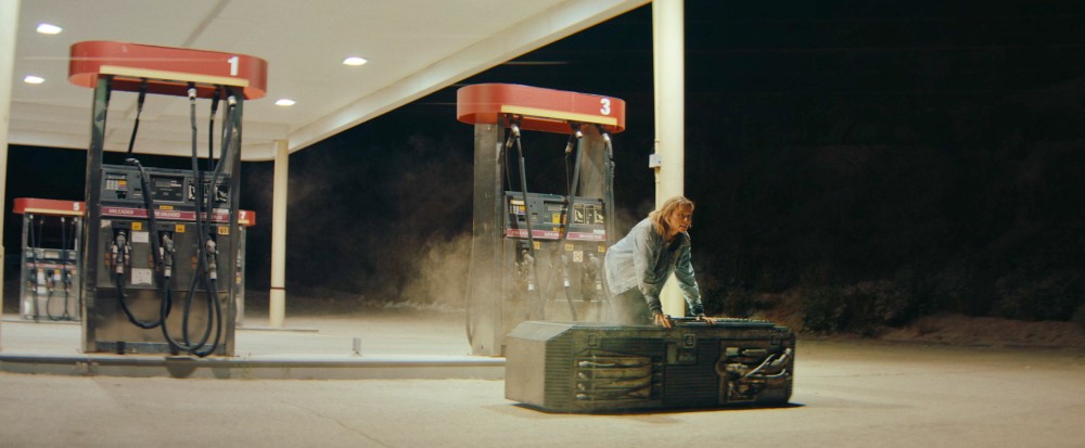 A petrol station at night. Where a car would be is instead a kind of steam punk metal coffin, with pipes and exhausts all over it. Steam is enveloping it and a tall, slim blonde woman is climbing out of it.