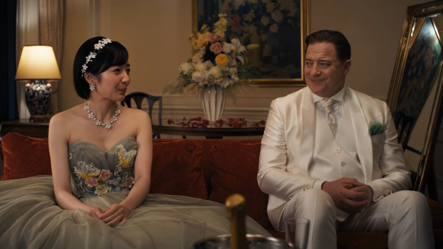 A young Japanese woman sat on the left hand side of a sofa. A middle aged, white American man sat on the right. She's wearing a grey wedding dress, he's wearing a cream wedding suit and both are smiling at each other awkwardly.