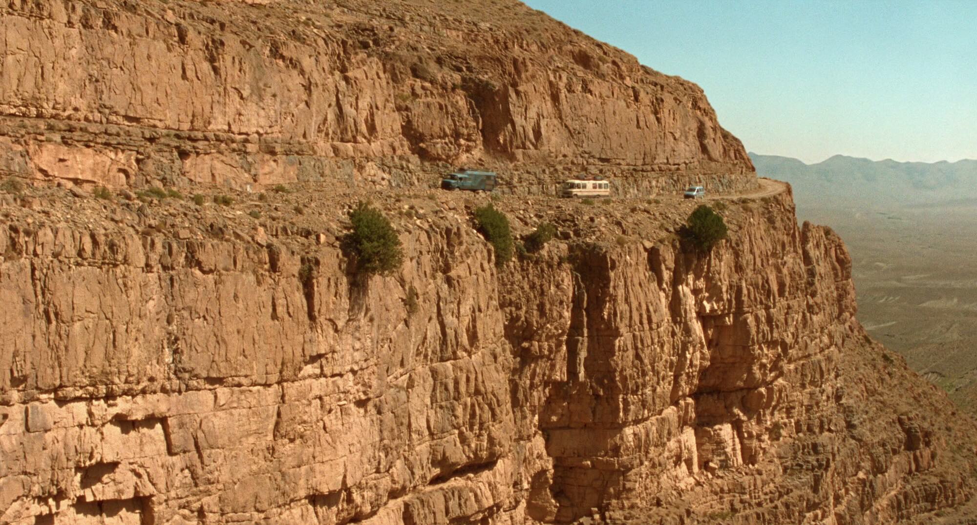 Wide shot of a vertiginous Moroccan mountain face. In the distance we can see two chunky buses, followed by a forlorn mini-van, are slowly edging along a narrow track impossibly etched into the side of the mountain.