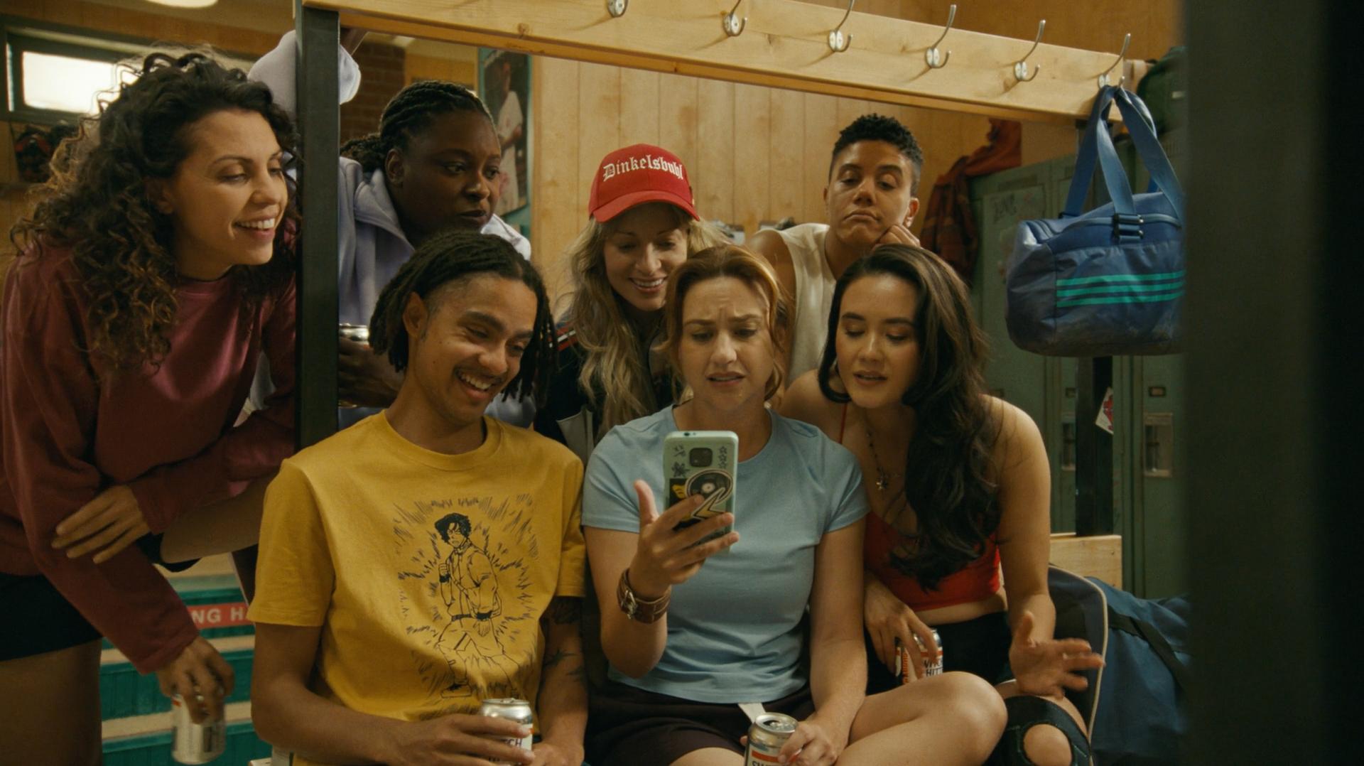 A group of six young women and one guy, in a locker room, all looking at the screen of a phone being held up the woman in the middle.