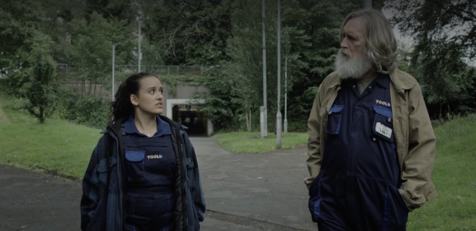 An older man with a long white beard and a young woman with dark hair walking along a path. Both are wearing dark blue coveralls, branded with the name of a DIY retailer.