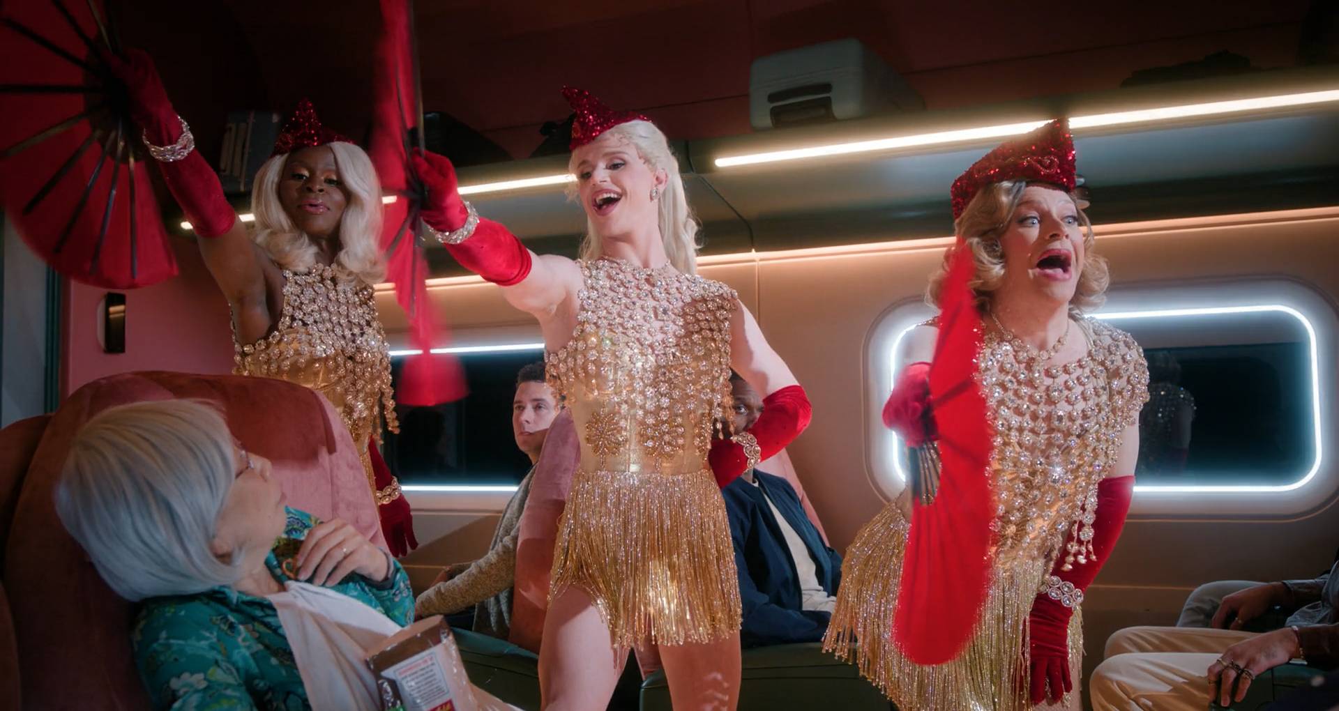 Three glamorous 'train stewardesses', dressed in white sequins and pearls, with Red hats and red fans, singing and dancing for the passengers, who look taken aback.