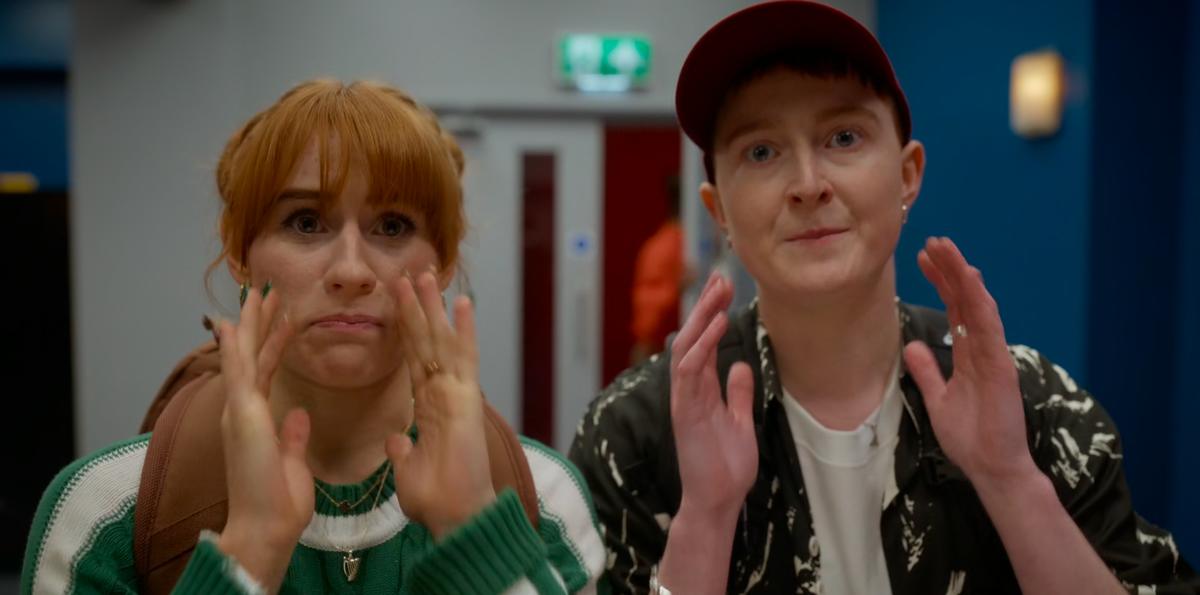 Head and shoulders shot of two young women, both looking into the camera. The one on the left has long, ginger hair. The one on the right has her hair completely covered by a baseball cap. Both have their hands raised to their faces in a look of shock.