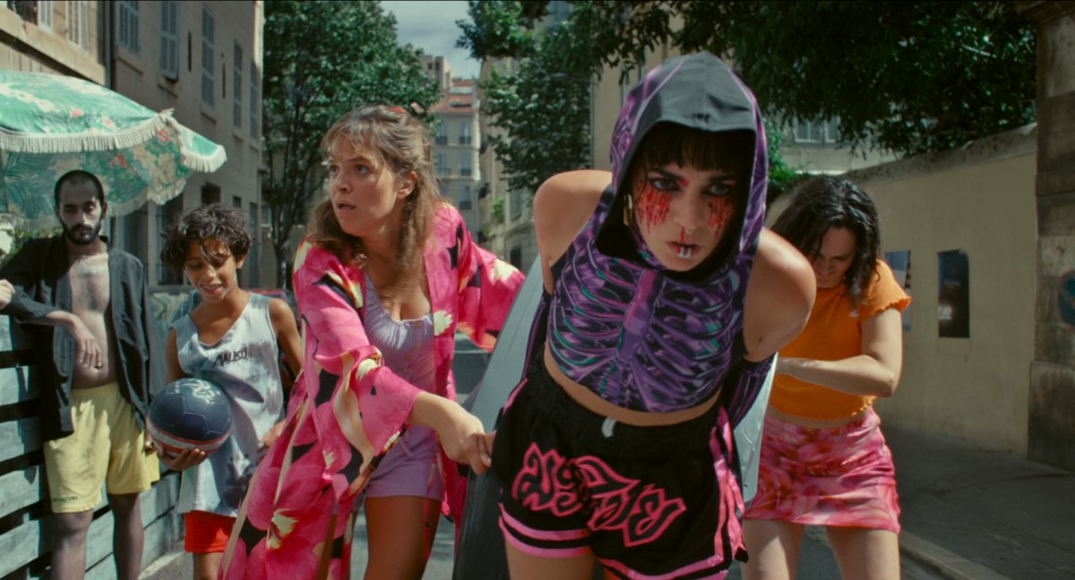 Three young women dragging a wheelie bin down a hot summer street in Marseille. The lead woman is wearing a matching purple outfit with a skeleton on it and red glitter mascara the makes it look like her eyes are bleeding. The other two are wearing floaty summer clothes. Watching them go past are an evidently amused young boy with a football and a non-plussed man who looks a bit like a zombie.