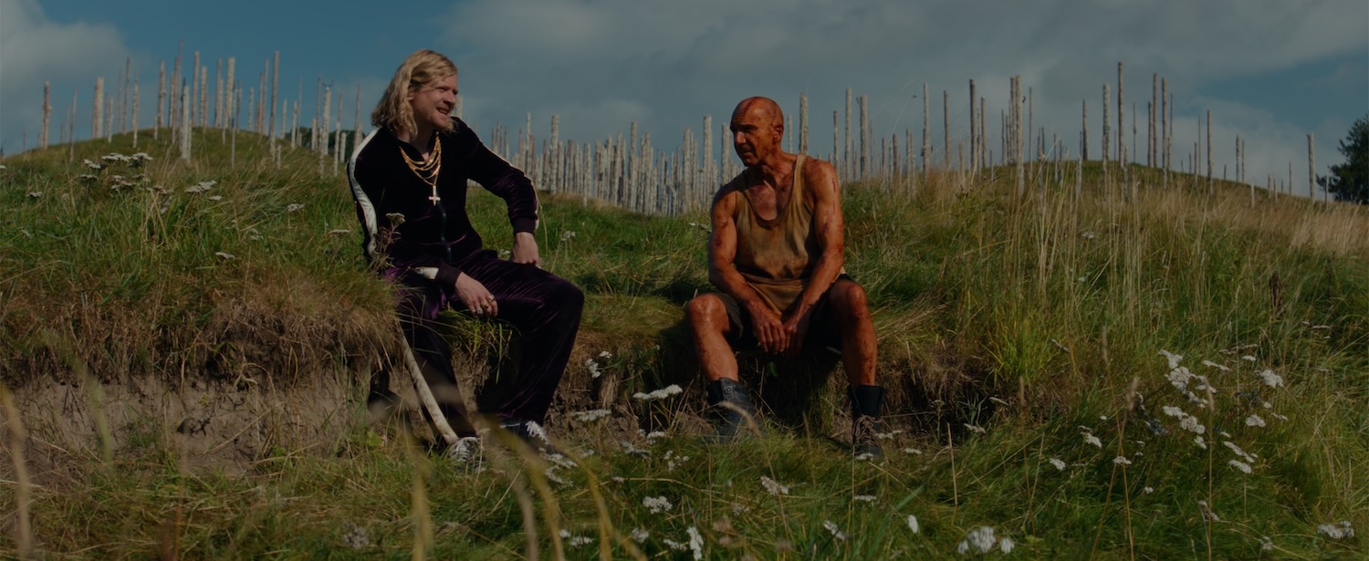 A field in England, in summer. Two men sit on the grass talking to each other. The one on the left is wearing a dark velour tracksuit and bunches of gold neck chains. He has long blonde hair. The one on the right is bald, painted red and looks tired. In the background are spires made of human skulls.