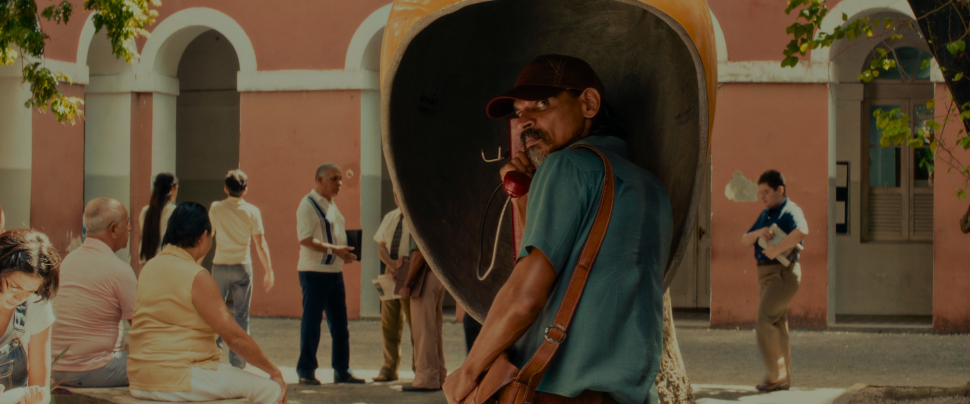 A skinny man with a greying goatee stands in a yellow phone kiosk in 70's Brazilian plaza. He's looking shiftily behind him.