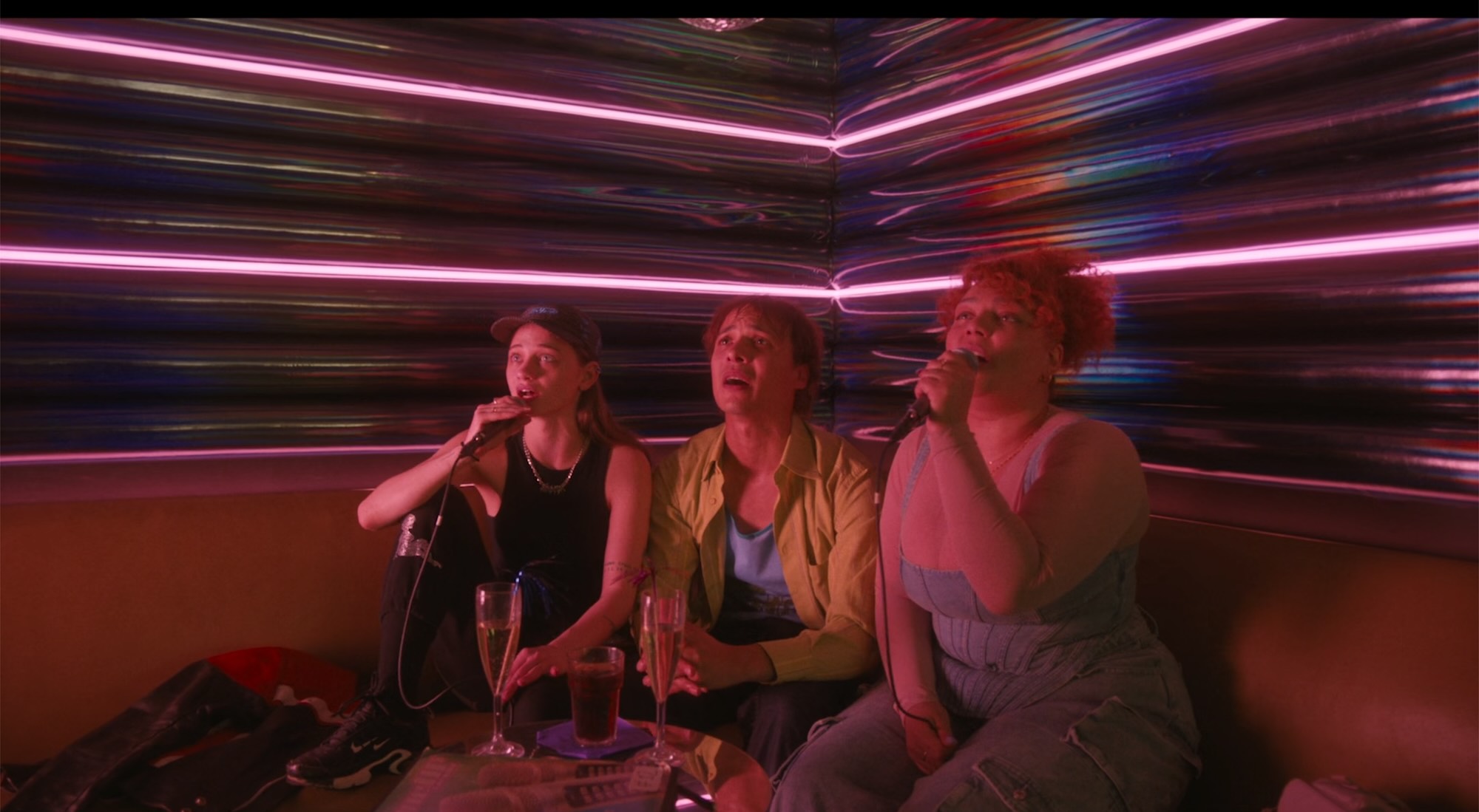 A neon drenched karaoke scene. Two young women flank a young man, all sat on a sofa singing into microphones.