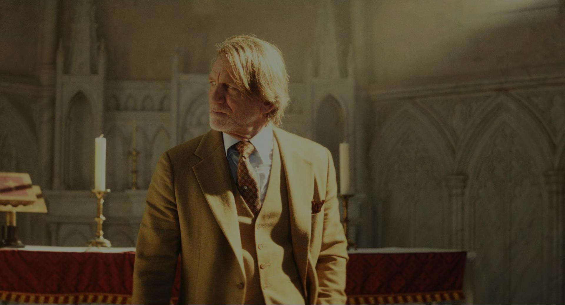 Daniel Craig in profile with straggly blond hair and a dapper sable suit. He's standing in a church with golden light shining on him.